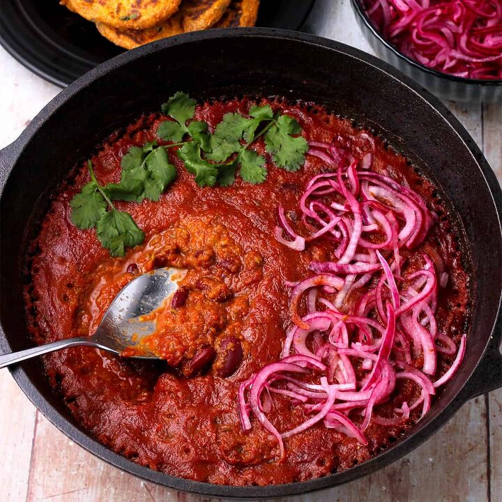 creamy rajma masala vegan kidney bean curry, A black cast iron pot is filled with rajma masala topped with pickled red onions and chopped cilantro