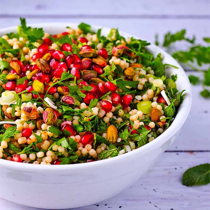 festive pomegranate pearl couscous salad, A white bowl filled with cooked Israeli couscous pomegranate herbs scallions and pistachios mixed in a white bowl