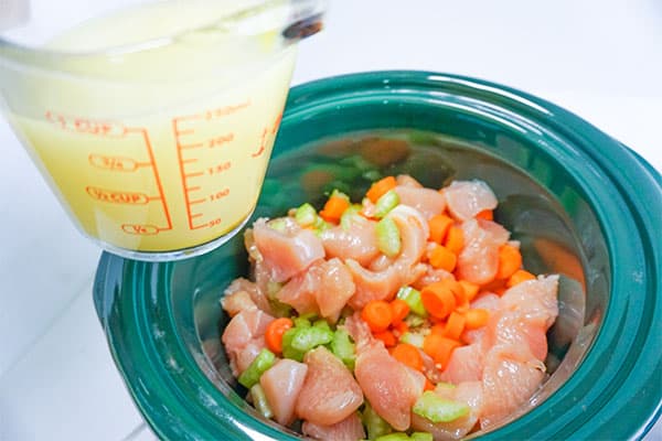 slow cooker chicken noodle soup, chicken broth in a glass measuring cup about to be added to raw cubed chicken and diced carrots and celery in a slow cooker