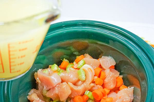 slow cooker chicken and dumplings, broth in a glass measuring cup above chicken celery carrot spices in a green slow cooker