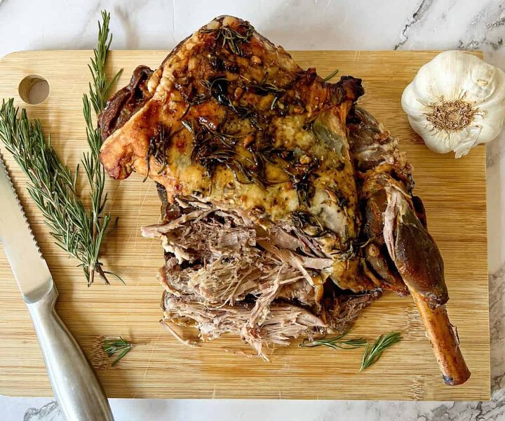 slow cooked leg of lamb in red wine, A carved leg of lamb on a chopping board with a knife There is a bulb of garlic and a sprig of rosemary also on the chopping board