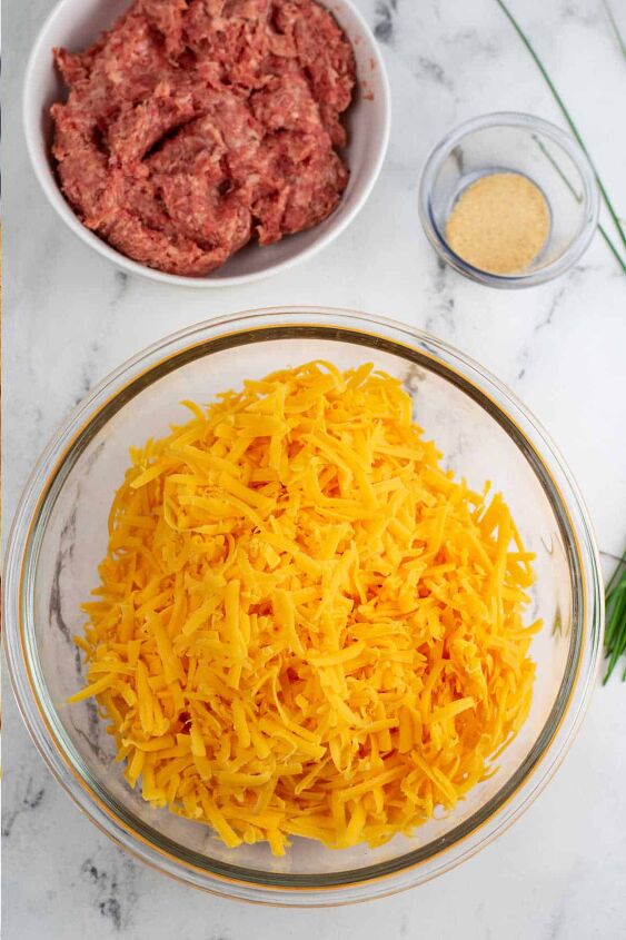 sausage and cheese balls, A large glass mixing bowl full of shredded cheddar cheese Next to it sits a bowl of sausage and a small bowl of garlic powder