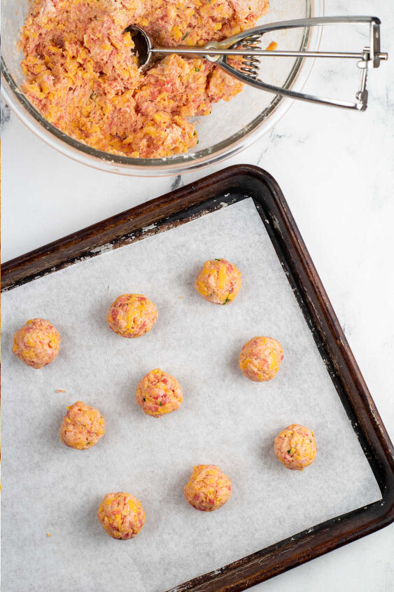 sausage and cheese balls, A glass mixing bowl with dough for sausage cheese balls in it There is a dough scoop in the bowl Next to it is a parchment lined baking sheet with balls of the sausage mixture on it