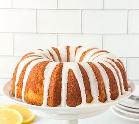 this lemon bundt cake recipe makes me happy, lemon bundt cake with white icing drizzle on top on a white cake stand Fresh sliced lemons in background Stack of small dishes in background