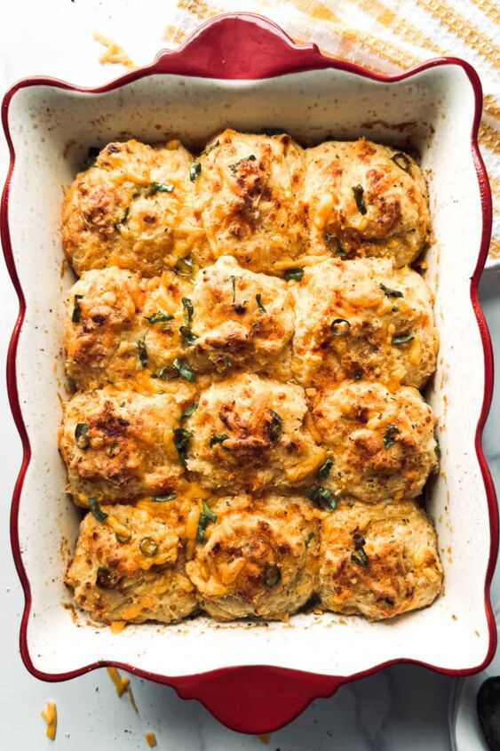 cheddar bay breakfast casserole with sausage and eggs, A red and white baking dish containing 12 golden brown biscuits garnished with herbs A yellow and white checkered cloth is partially visible in the background