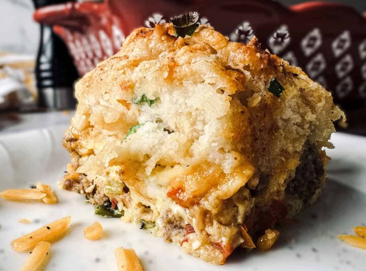 cheddar bay breakfast casserole with sausage and eggs, Close up of a savory casserole slice with visible chunks of vegetables meat and herbs The dish appears to be baked with a golden brown top and a fluffy moist interior served on a white plate with scattered bits of cheese in the foreground