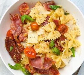 creamy bow tie pasta salad with crispy pancetta, A bowl of bowtie pasta topped with crispy prosciutto cherry tomatoes shaved parmesan and fresh basil leaves The dish is seasoned with black pepper and presented on a white plate with a light blue background