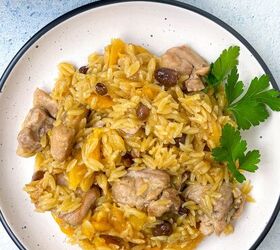 chicken orzo bake one pan family dinner, A plate of orzo with chunks of meat raisins and herbs garnished with parsley The dish is served on a white plate with a black rim set against a light blue background
