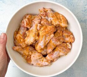garlic butter chicken wings oven baked, A hand holds a white bowl brimming with raw chicken wings marinated in a reddish garlic butter seasoning all set against a light blue background