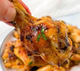 garlic butter chicken wings oven baked, A hand holding a glazed grilled chicken wing garnished with herbs A plate of garlic butter chicken wings is visible in the background The glaze appears shiny highlighting the crispy texture of the wing