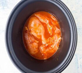 how to make shredded chicken in crockpot, A black slow cooker bowl containing raw chicken pieces covered in a red marinade or sauce set against a light blue and white background