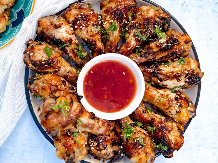 easy soy garlic chicken wings recipe, A plate of grilled soy garlic chicken drumsticks artfully arranged in a circle around a small bowl of red dipping sauce The chicken is garnished with chopped herbs and sesame seeds set against a light blue background