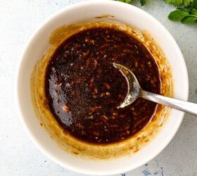 easy soy garlic chicken wings recipe, A white bowl filled with a dark thick sauce for soy garlic chicken wings featuring visible spices and a glossy texture A spoon rests inside the bowl Fresh cilantro leaves are visible in the top right corner on a light textured surface