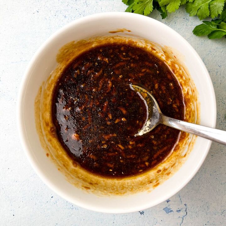 easy soy garlic chicken wings recipe, A white bowl filled with a dark thick sauce for soy garlic chicken wings featuring visible spices and a glossy texture A spoon rests inside the bowl Fresh cilantro leaves are visible in the top right corner on a light textured surface
