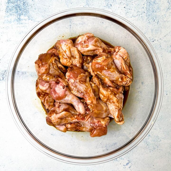 easy soy garlic chicken wings recipe, A glass bowl filled with marinated raw chicken wings rests on a light gray surface The marinade with hints of soy and garlic gives the wings a glossy reddish brown appearance