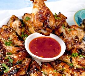 easy soy garlic chicken wings recipe, A plate of soy garlic chicken wings garnished with sesame seeds and herbs A hand holds one wing dipped in a small bowl of red sauce at the center The wings are arranged in a circular pattern around the sauce showcasing their irresistible glaze