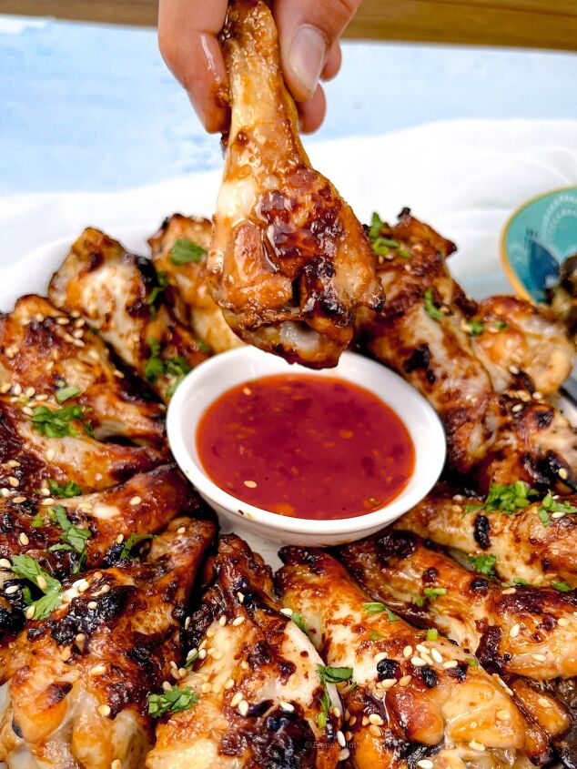 easy soy garlic chicken wings recipe, A plate of soy garlic chicken wings garnished with sesame seeds and herbs A hand holds one wing dipped in a small bowl of red sauce at the center The wings are arranged in a circular pattern around the sauce showcasing their irresistible glaze