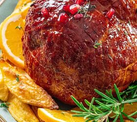 the 3 ingredient ham glaze recipe that changes everything, The image shows roasted ham topped with a glaze garnished with pomegranate seeds It is surrounded by orange slices and sprigs of rosemary Roasted potatoes are placed alongside the ham on a green plate