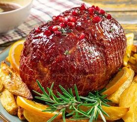 the 3 ingredient ham glaze recipe that changes everything, The image shows a glazed ham topped with red berries Surrounding the ham are orange slices and roasted potatoes garnished with sprigs of rosemary