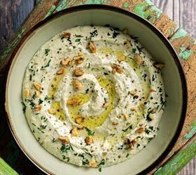 A bowl of creamy hummus garnished with olive oil chopped herbs and nuts sits on a weathered green wooden chair The rustic background enhances the homemade and natural appearance of the dish