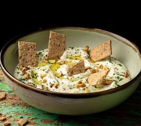 A bowl of creamy white dip garnished with olive oil herbs and nuts topped with several rectangular whole grain crackers It is set against a rustic green surface with cracker crumbs scattered around