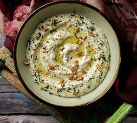 A bowl of creamy dip garnished with chopped walnuts herbs and a drizzle of olive oil The bowl is set on a rustic wooden surface with a rich textured maroon cloth partially visible beside it