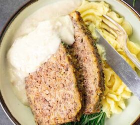 homemade meatloaf with creamy onion gravy, A plate with slices of meatloaf topped with gravy served with mashed potatoes and garnished with rosemary A fork and knife are placed beside the dish and a jug of additional gravy is visible in the background