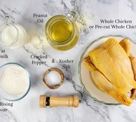 how to make buttermilk fried chicken, A top view of cooking ingredients including a whole chicken on a plate a glass of buttermilk a jug of peanut oil a bowl of self rising flour cracked pepper kosher salt and a pepper grinder on a marble surface Labels identify each item