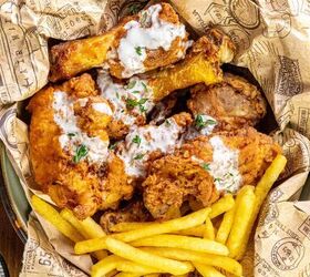 how to make buttermilk fried chicken, A basket lined with printed paper holds crispy fried chicken topped with creamy sauce and garnished with herbs Golden fries are served on the side A small dish of spices is visible in the background on a wooden table