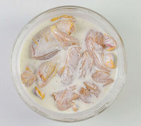 how to make buttermilk fried chicken, A glass bowl filled with raw chicken pieces marinating in a creamy white yogurt mixture atop a light gray surface