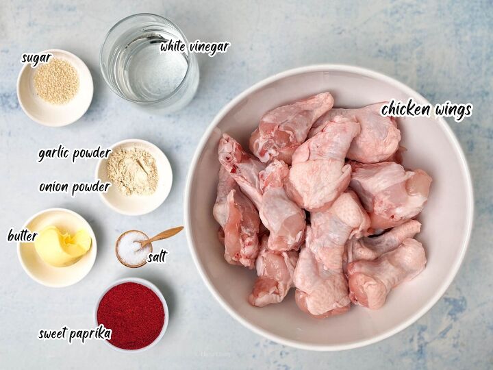 crispy salt and vinegar chicken wings recipe, A top view image of raw chicken wings in a bowl surrounded by small dishes containing white vinegar sugar garlic powder onion powder butter salt and sweet paprika Each ingredient is labeled in text The background is a light blue surface