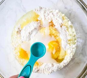 pina colada cupcakes, A hand holding a measuring spoon over a glass bowl full of igredients for making pina colada cupcakes