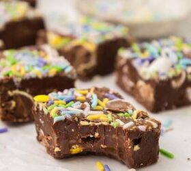 easter fudge, Close up of chocolate fudge pieces with colorful sprinkles and chocolate eggs on top arranged on white parchment paper One piece has a bite taken out revealing a rich soft texture A small dish is blurred in the background