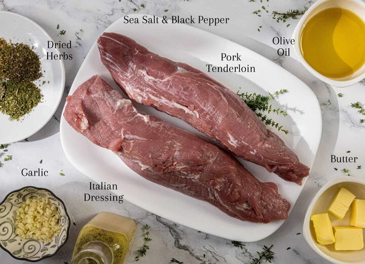 southern oven roasted pork tenderloin with herb crust, Two raw pork tenderloins on a white plate surrounded by bowls of olive oil butter cubes chopped garlic Italian dressing dried herbs and sea salt with black pepper all placed on a marble surface with scattered thyme sprigs