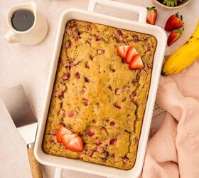 strawberry banana pancake casserole, A rectangular strawberry banana pancake casserole in a white baking dish is on a table Sliced strawberries garnish the top Side items include a pink cloth a cake server a banana a cup of coffee and a bowl of fresh strawberries