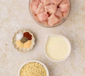 oven baked chicken nuggets, A top down view of cubed raw chicken in a bowl surrounded by small bowls containing breadcrumbs melted butter and a mix of spices including salt pepper and paprika all on a light colored surface