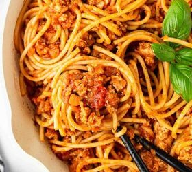 ground turkey with spaghetti, A close up of spaghetti with meat sauce in a white pan garnished with fresh basil Black spoons are holding up a twirl of pasta A striped cloth is partially visible in the corner