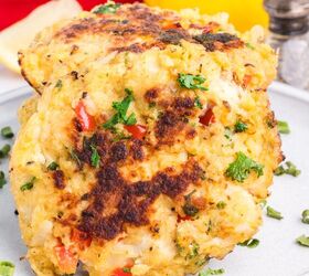 classic crab cake recipe little filler, Two golden brown crab cakes with chopped herbs and red pepper are stacked on a plate with a lemon red bell pepper and salt shaker in the background