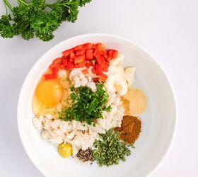 classic crab cake recipe little filler, A white bowl containing chopped red pepper egg mayonnaise crab meat parsley spices and seasonings sits on a white surface with fresh parsley and a red bell pepper nearby