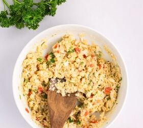classic crab cake recipe little filler, A bowl of mixed ingredients with chopped parsley and red bell pepper stirred with a wooden spoon sits on a white surface alongside fresh parsley and a red bell pepper