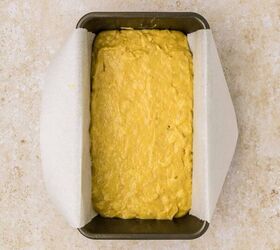 cake mix banana bread, A rectangular metal loaf pan lined with parchment paper is filled with unbaked cake mix banana bread batter The pan is placed on a beige textured surface