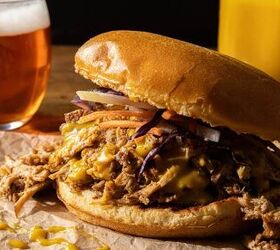 A pulled pork sandwich topped with melted cheese coleslaw and sauce on a bun served on parchment paper In the background there is a glass of beer and a glass of orange juice