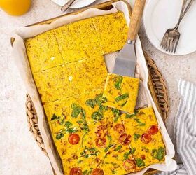sheet pan eggs, A baking tray with parchment paper holds a sliced baked eggs with vegetables A spatula lifts a piece Plates forks orange juice and a bowl of shredded cheese are nearby on a light countertop