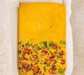 sheet pan eggs, Rectangular baking pan lined with parchment paper filled with an uncooked egg mixture The bottom half has visible spinach tomatoes and cheese while the top half is only yellow liquid
