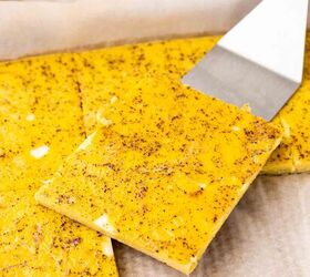 sheet pan eggs, A metal spatula lifts a square slice of baked egg from a parchment lined baking sheet The egg squares are yellow with visible seasoning and more pieces are on the tray and a white plate in the background