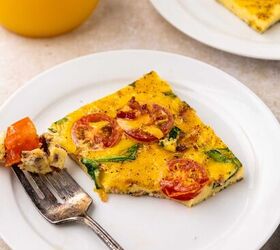 sheet pan eggs, A slice of eggs with vegetables on top with cherry tomatoes and greens on a white plate with a fork holding a bite A glass of orange juice and another plate with more sheet pan eggs are in the background