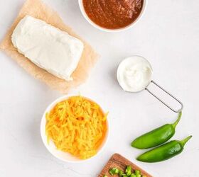 salsa and cream cheese dip, Ingredients on a white surface a block of cream cheese on parchment a bowl of salsa a small cup of sour cream shredded cheddar cheese in a bowl two whole jalape os and chopped jalape os on a cutting board with a knife