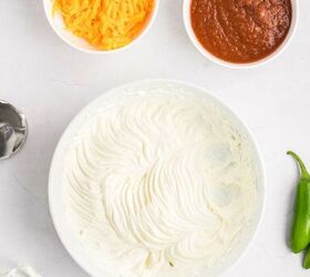 salsa and cream cheese dip, A bowl of whipped cream cheese and sour cream sits beside a small bowl of shredded cheddar cheese and a bowl of salsa Part of a hand mixer and beaters are visible along with a jalape o on a white surface