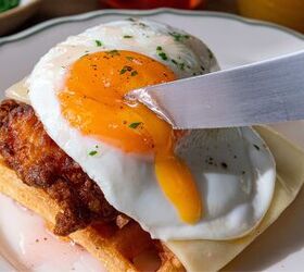A knife cuts into a runny fried egg yolk resting on a piece of crispy fried chicken atop a waffle The dish is garnished with a sprinkle of herbs and placed on a white plate with a green rim