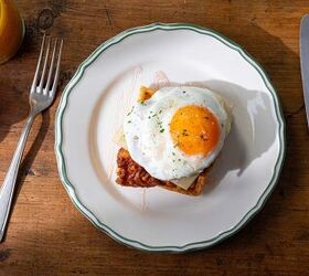 A breakfast plate with a croque madame on a wooden table The sandwich consists of toasted bread ham and melted cheese topped with a fried egg A fork a knife a small jar of jam and a glass of orange juice are nearby
